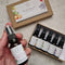 A hand holds a small amber pillow spray bottle, with the matching kraft gift box and an open tray of five similar bottles in the background on a light stone surface.