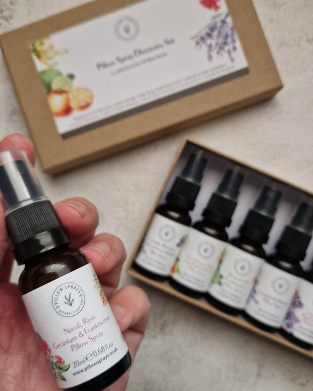 A hand holds a small amber pillow spray bottle, with the matching kraft gift box and an open tray of five similar bottles in the background on a light stone surface.