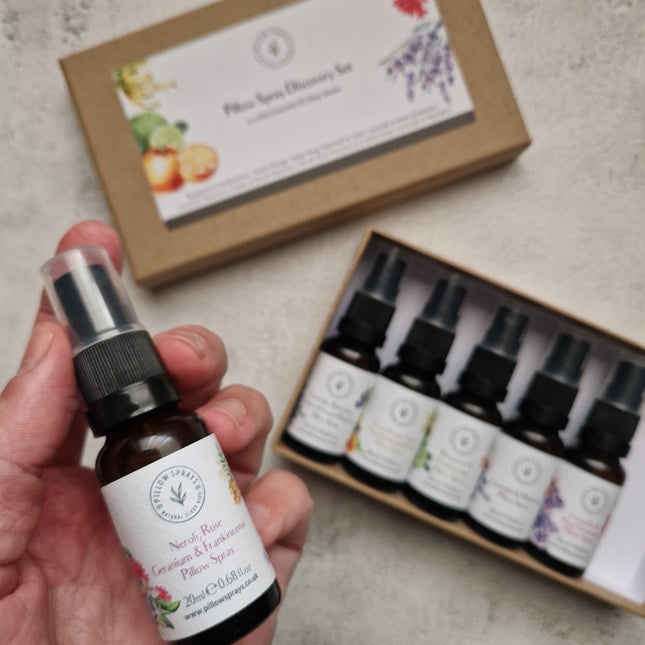 A hand holds a small amber pillow spray bottle, with the matching kraft gift box and an open tray of five similar bottles in the background on a light stone surface.