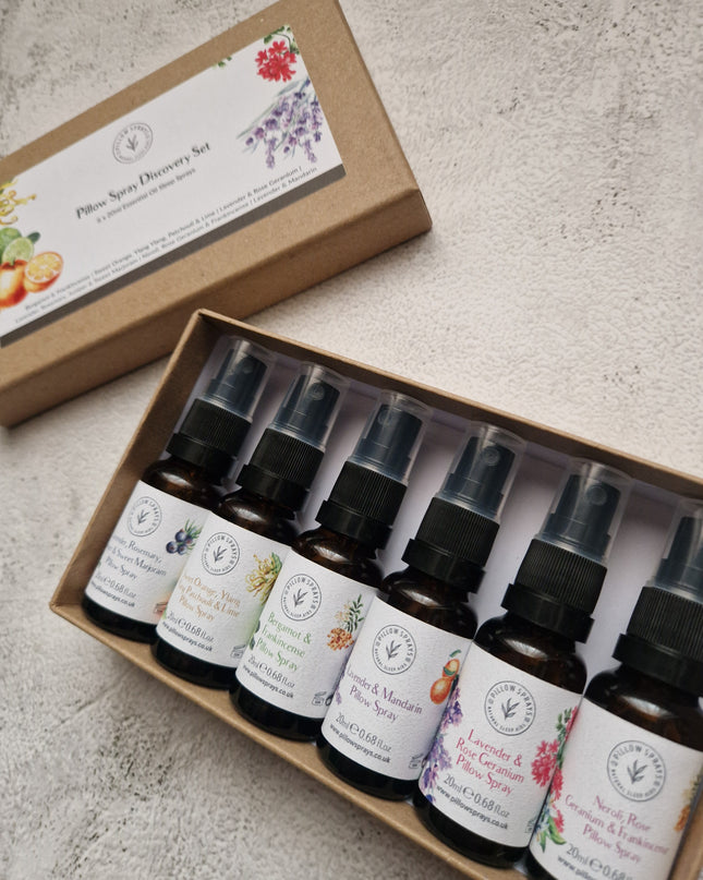 A boxed set of six small amber glass pillow spray bottles, arranged in an open kraft paper tray beside the matching lid on a light textured surface.