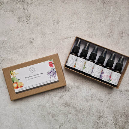 A boxed set of six essential oil pillow spray bottles displayed on a textured light gray surface, with the lid placed beside the open tray. The set includes botanical illustrations on the labels and packaging.