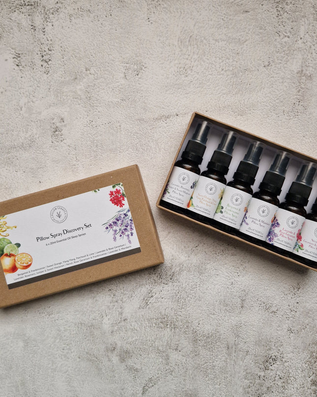 A boxed set of six essential oil pillow spray bottles displayed on a textured light gray surface, with the lid placed beside the open tray. The set includes botanical illustrations on the labels and packaging.