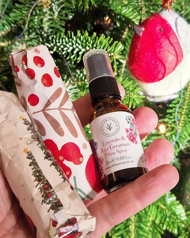 Hand holding a small bottle of essential oil pillow spray with decorative paper against a Christmas tree background